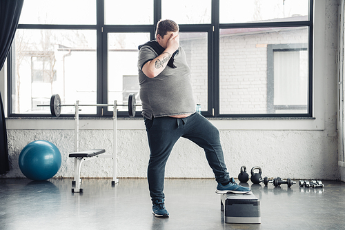overweight man with towel covering face with hand while training on step platform at sports center