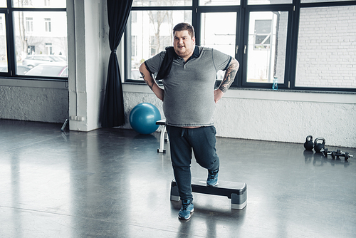 overweight man in grey t-shirt with towel training on step platform at sports center