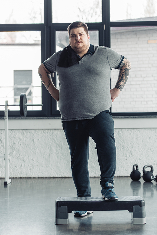 overweight tattooed man with towel Looking At Camera while training on step platform at sports center