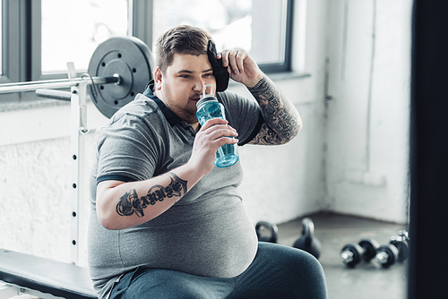 overweight tattooed man drinking water from sport bottle and wiping face with towel at gym