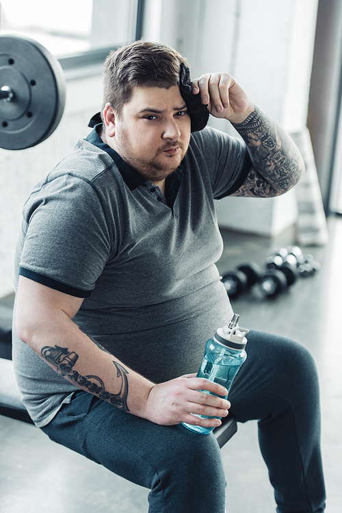overweight tattooed man holding sport bottle,  and wiping face with towel at gym