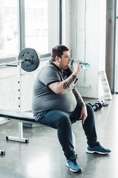 overweight tattooed man sitting on bench and drinking water from sport bottle at gym
