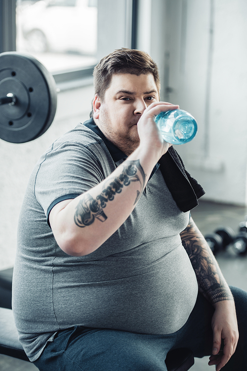 tired overweight tattooed man drinking water from sport bottle at gym