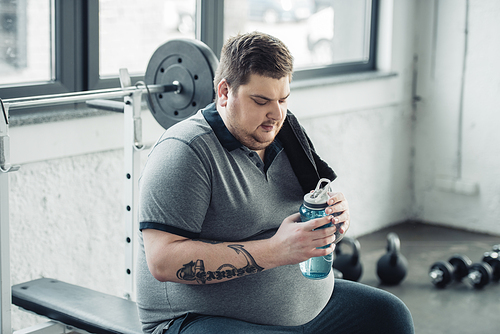 overweight tattooed man sitting on bench and holding sport bottle at gym