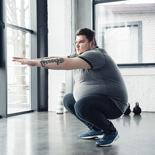 overweight tattooed man doing squats at sports center