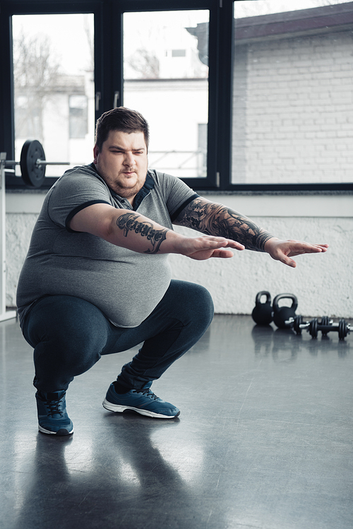 overweight tattooed man doing squats at sports center