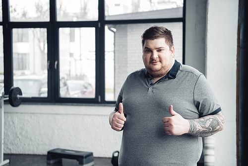 overweight tattooed man Looking At Camera and showing thumbs up at gym with copy space