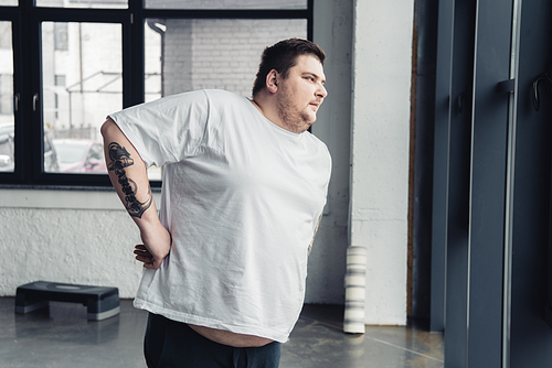obese tattooed man in white t-shirt doing stretching exercise at sports center