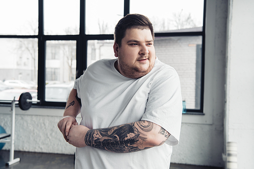 overweight tattooed man in white t-shirt stretching arms at gym