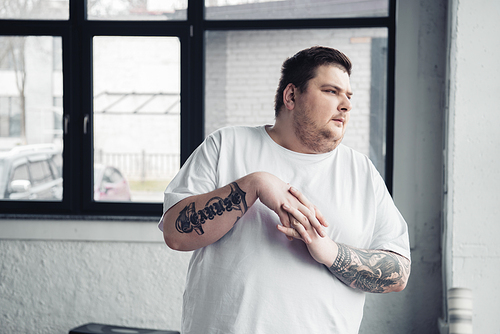 overweight tattooed man in white t-shirt stretching fingers at gym