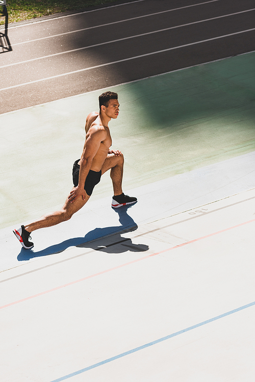 shirtless mixed race sportsman in black sneakers stretching at stadium