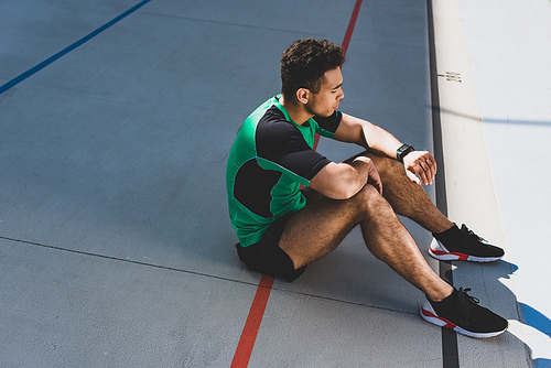 handsome mixed race sportsman sitting on running track and looking at smartwatch