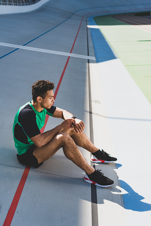 handsome mixed race sportsman sitting on running track at stadium and looking at smartwatch