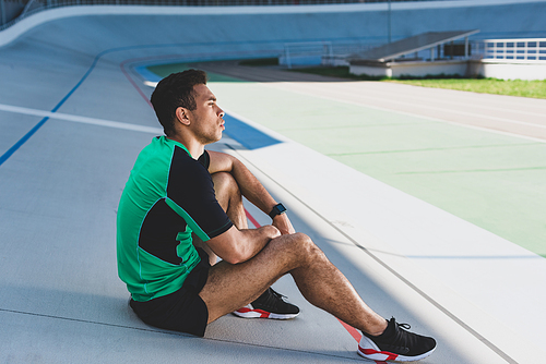 mixed race sportsman sitting on running track and looking away at stadium