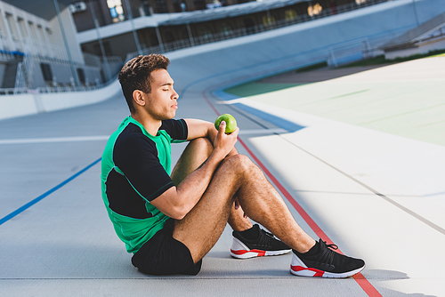 side view of mixed race sportsman sitting in relax manner on running track and looking at green apple