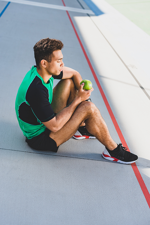 mixed race sportsman sitting on running track and holding green apple