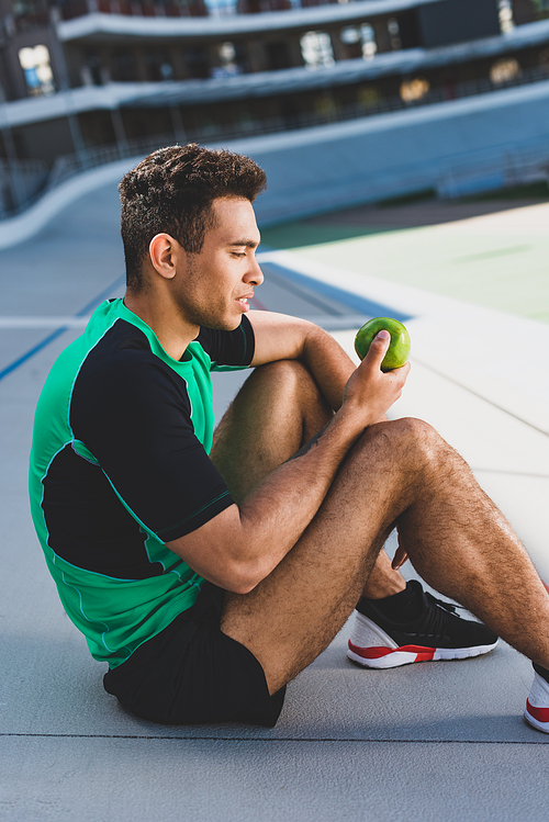 side view of mixed race sportsman sitting on running track and looking at green apple