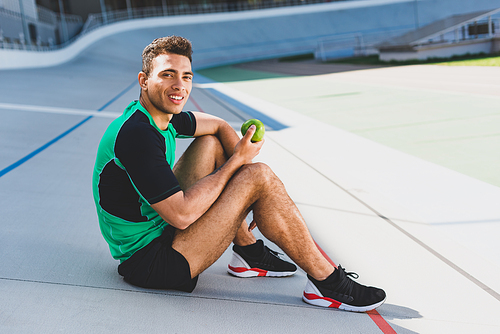 mixed race sportsman sitting on running track at stadium,  and holding green apple