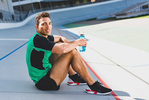 mixed race sportsman sitting on running track at stadium,  and holding bottle with water