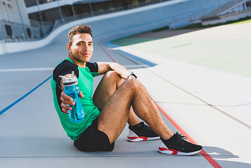 mixed race sportsman sitting on running track at stadium, looking and presenting bottle with water at camera