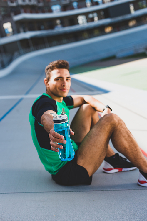 selective focus of mixed race sportsman sitting on running track at stadium, looking and presenting bottle with water at camera