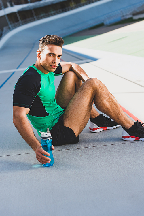 mixed race sportsman sitting on running track, seriously  and holding bottle with water