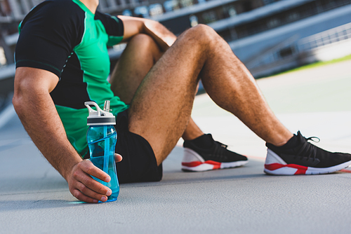 cropped view of sportsman sitting on running track and holding bottle with water