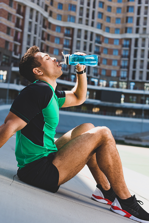 mixed race sportsman sitting on running track at stadium and drinking water from blue bottle