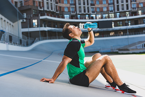 side view of mixed race sportsman sitting on running track at stadium and drinking water from blue bottle