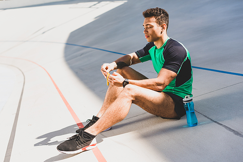 mixed race sportsman sitting on running track near blue bottle and opening chocolate bar