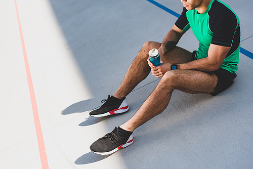 cropped view of sportsman sitting on running track and holding blue bottle with water