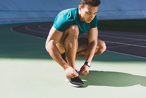 mixed race sportsman lacing up sneakers at stadium in sunny weather