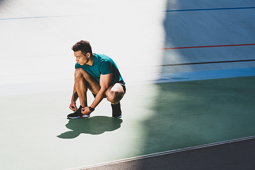 mixed race sportsman lacing up sneakers at stadium, standing on green floor