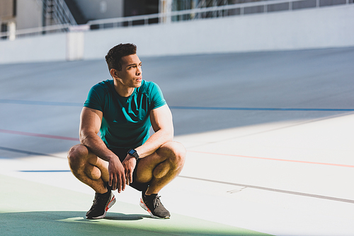front view of mixed race sportsman squatting and looking away at stadium in sunlight
