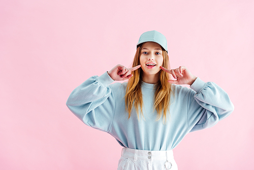 pretty teenage girl in cap pointing with fingers at smile isolated on pink