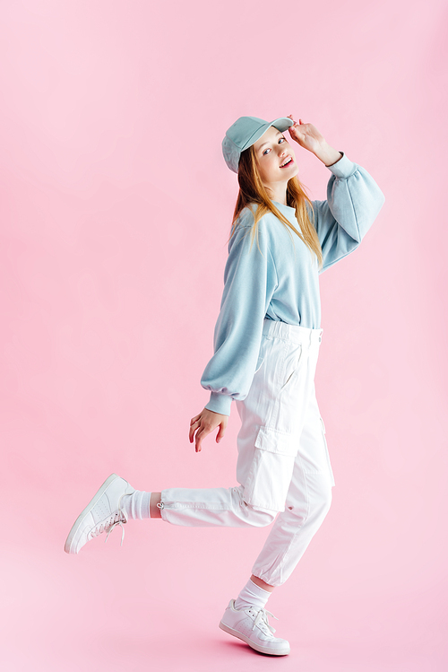 full length view of smiling pretty teenage girl in cap on pink