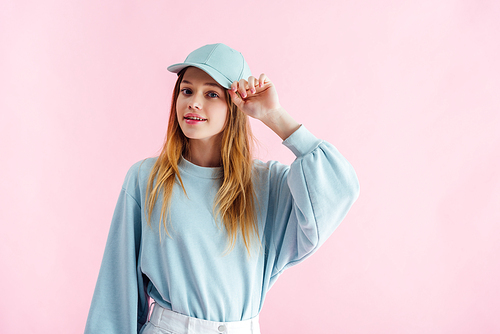 smiling pretty teenage girl touching cap isolated on pink