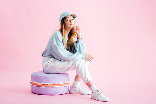 pretty teenage girl in cap sitting on decorative macaroon on pink