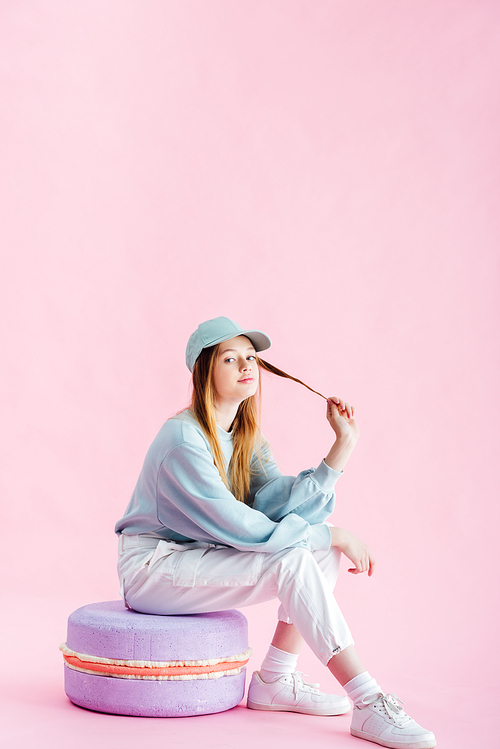 pretty teenage girl in cap touching hair while sitting on decorative macaroon isolated on pink