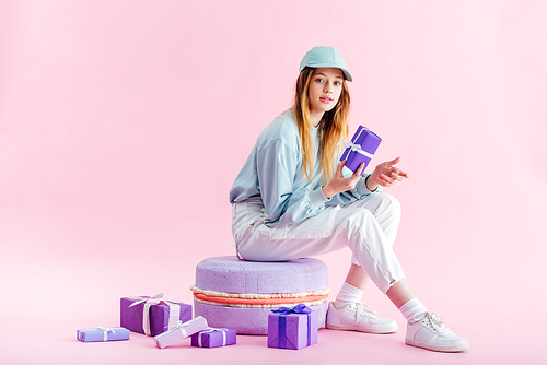 teenage girl in cap sitting on decorative macaroon near presents on pink