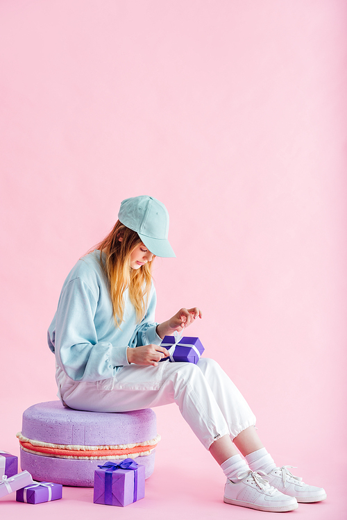 pretty teenage girl in cap sitting on decorative macaroon and unpacking presents on pink