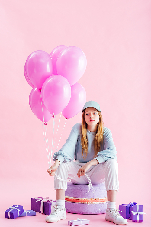 teenage girl in cap sitting on decorative macaroon with balloons near gifts on pink