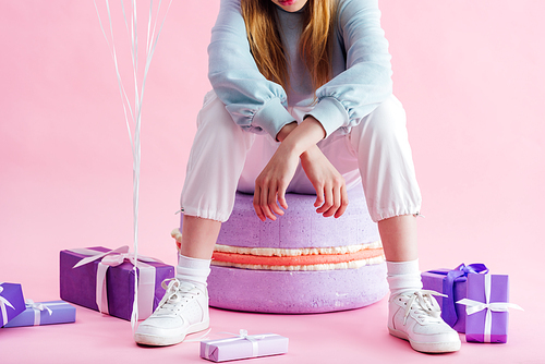 cropped view of teenage girl sitting on decorative macaroon near presents on pink