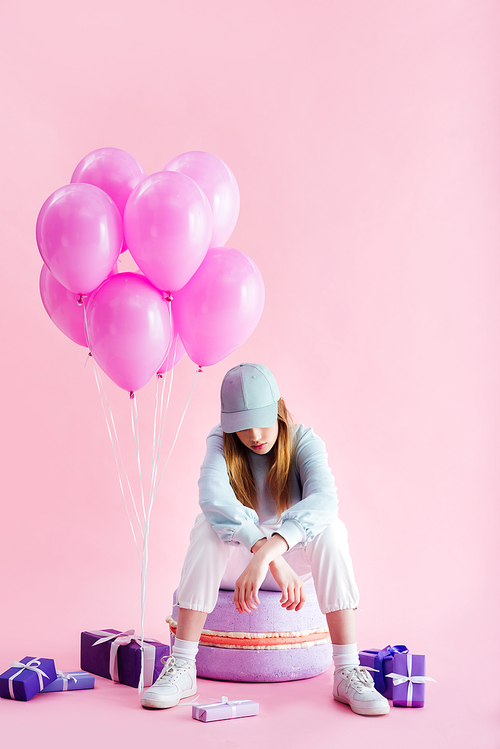 teenage girl in cap sitting on decorative macaroon near presents and balloons on pink