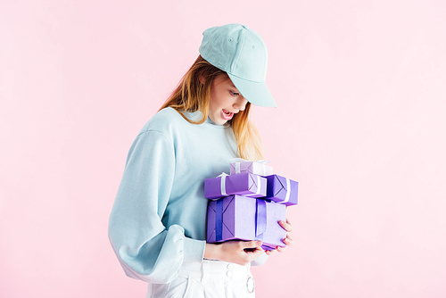 excited pretty teenage girl in cap looking at purple gifts isolated on pink