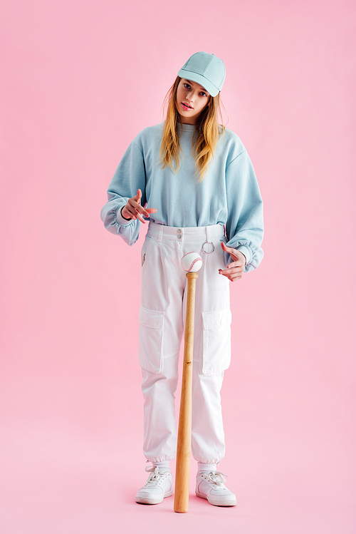 full length view of pretty teenage girl in cap near baseball ball on bat on pink