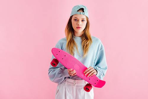 pretty teenage girl in cap holding penny board isolated on pink