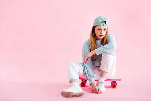 pretty teenage girl in cap sitting on penny board on pink