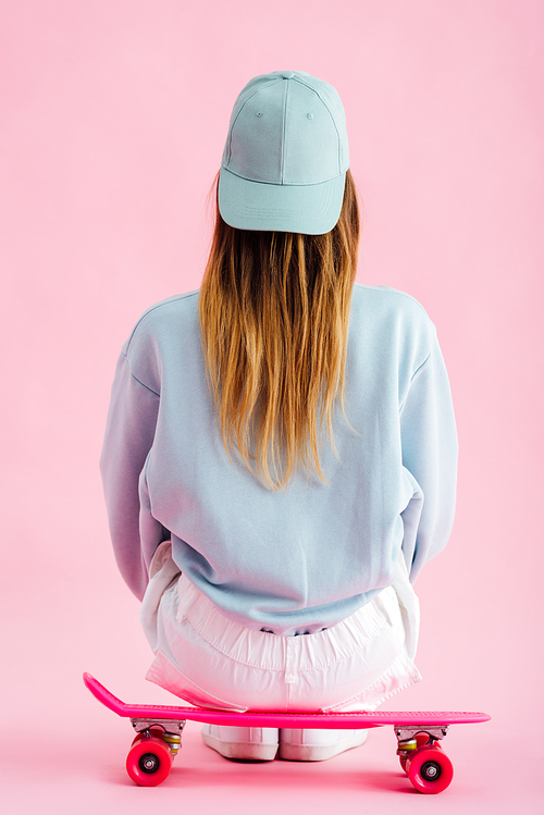 back view of teenage girl in cap sitting on penny board on pink