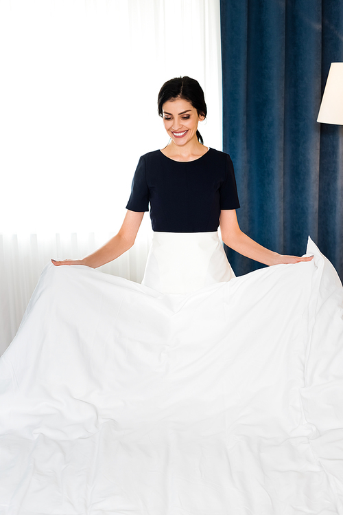 happy housemaid holding white bed sheet in hotel room